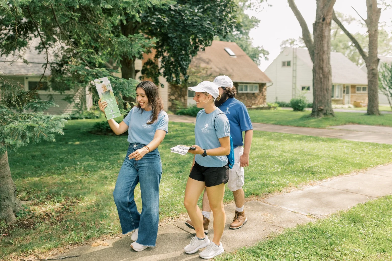 Four people canvassing with SBA Pro-Life America walk along a suburban sidewalk in Toledo, Ohio. Two women up front smile and talk, holding flyers, while two men follow behind. All are casually dressed, wearing hats. Houses line the background.