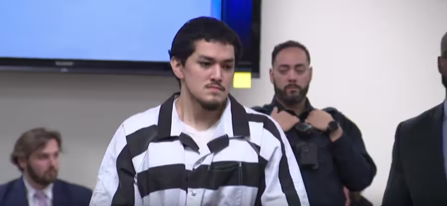 A man in a black-and-white striped prison uniform stands in a courtroom, with seated people and security personnel visible in the background.