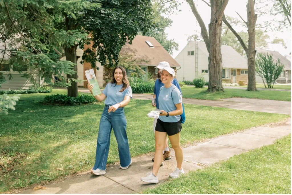 Three people walk along a suburban sidewalk in Toledo, Ohio, smiling and holding flyers as they canvass for SBA Pro-Life America on a sunny day, with houses and trees lining the background.