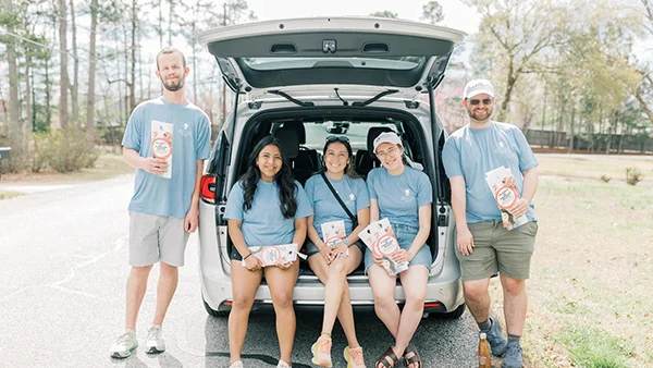 Five people wearing matching light blue shirts sit and stand by an open car trunk on a sunny day, each holding a bag of snacks—getting ready to start a fundraiser. Trees and greenery form the backdrop for this cheerful fundraising moment.