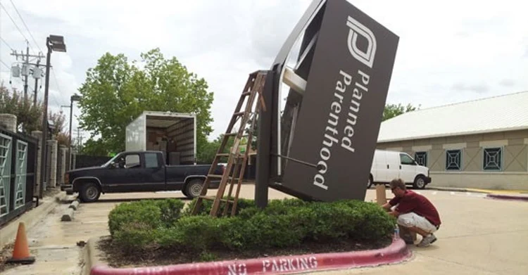 A large Planned Parenthood sign is tilted and partially propped up by a wooden ladder. A person crouches nearby with tools, and two parked vehicles are visible in the background. The curb reads NO PARKING.