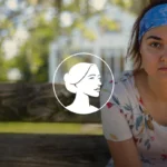 A woman wearing a blue headband and floral shirt sits on a wooden bench outdoors, looking slightly to the side with a neutral expression—her gaze thoughtful, perhaps reflecting on issues like the recent abortion drug lawsuit. The background is blurred with green trees and sunlight.