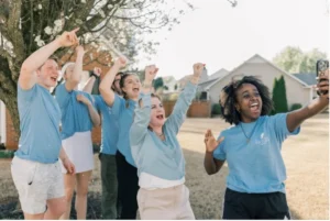 Six people outdoors celebrate and smile while taking a group selfie; they wear blue shirts promoting the Pro-Life cause and stand near a tree and houses on a sunny day.