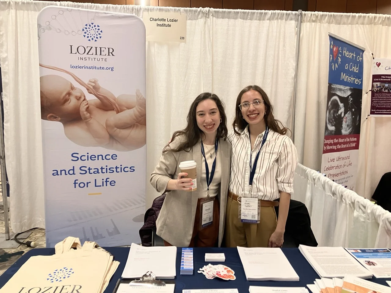 Two women, who appear to be interns, stand smiling behind a Lozier Institute booth with pamphlets, tote bags, and stickers. A banner shows a baby and reads “Science and Statistics for Life.” Both women wear badges and lanyards at a conference hosted by SBA for Pro-Life Leaders.