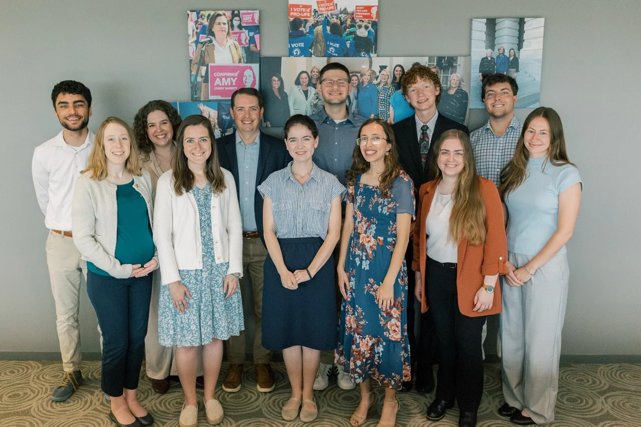 A group of thirteen SBA Interns pose and smile indoors in front of a gray wall adorned with colorful campaign posters and photos reflecting the current political climate. The group is dressed in business casual attire.