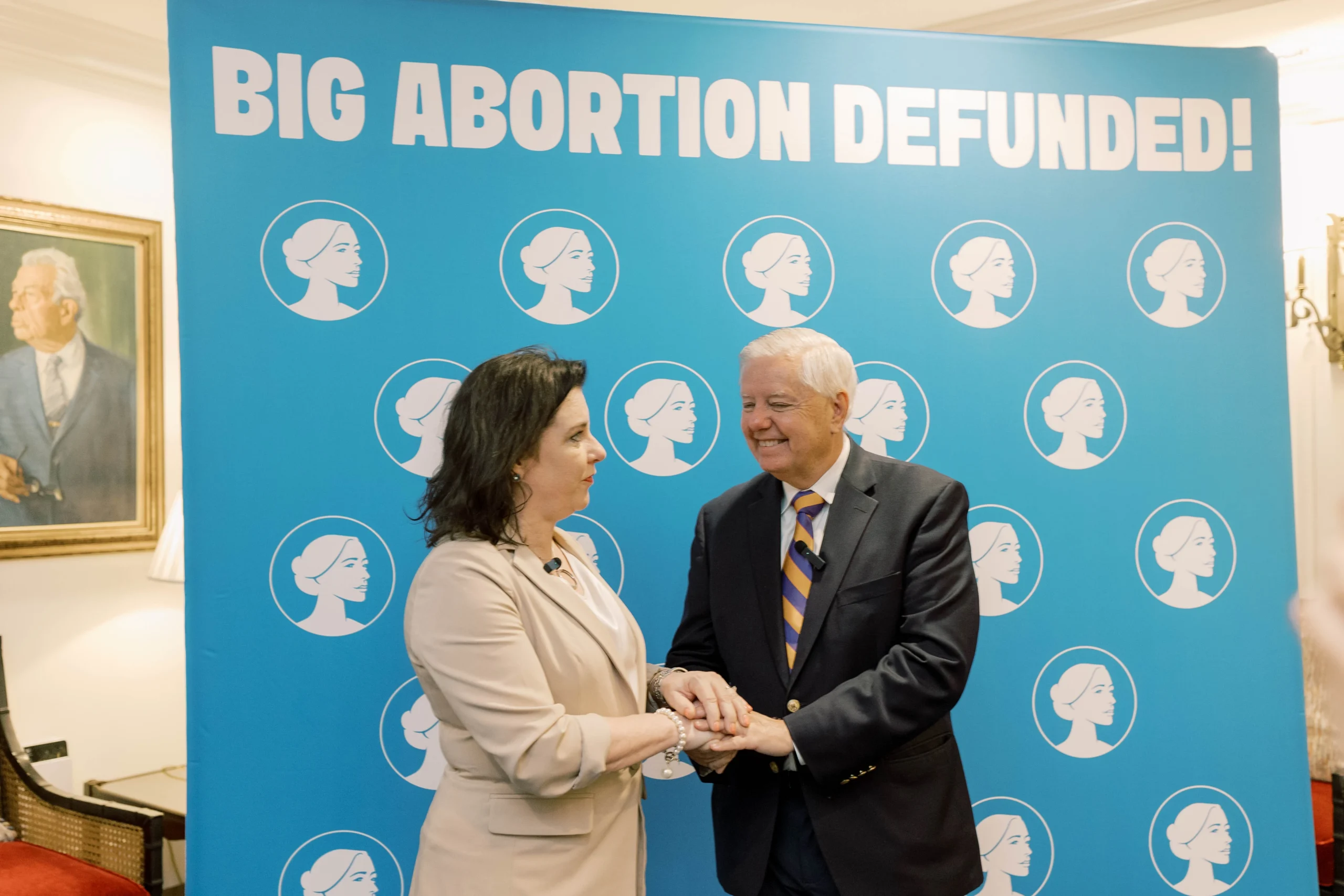 Two people shake hands in front of a blue banner that reads BIG ABORTION DEFUNDED! with repeated white profile icons of a womans face. A portrait hangs on the wall to the left.