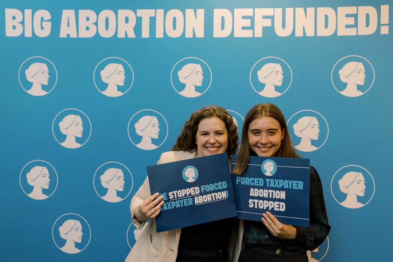 Two smiling interns hold blue signs reading STOPPED FORCED TAXPAYER ABORTION in front of a blue backdrop that says BIG ABORTION DEFUNDED! and features a repeated white logo of a woman’s profile, highlighting their pride as emerging pro-life leaders with SBA.