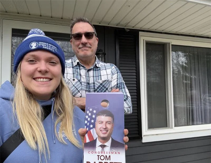 A young woman in a blue hat and a man stand outside a house. The woman, a student canvasser, holds a door hanger with a photo of a politician and an American flag, labeled CONGRESSMAN TOM.
