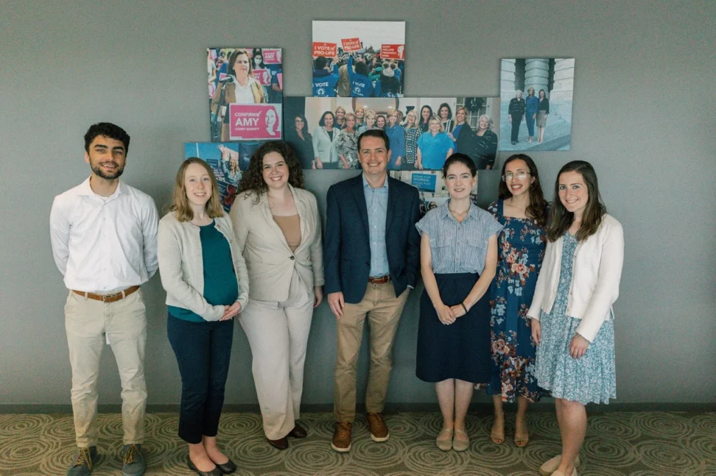 Seven people stand in a row, smiling, in front of a gray wall decorated with a collage of colorful photos and posters. Dressed in business casual attire, these SBA interns and Pro-Life Leaders exude enthusiasm and camaraderie.