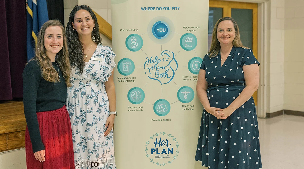Three women stand smiling beside a Her PLAN banner that outlines various support services, including care coordination, legal aid, recovery, financial assistance, and health and well-being. These Pro-Life Leaders exemplify compassion and dedication in supporting their community.