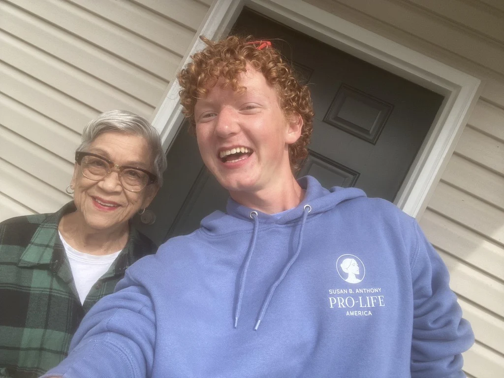 A young person in a blue hoodie and an older woman with glasses—possibly SBA Interns from different stages of life—stand smiling together in front of a beige house with a black door.