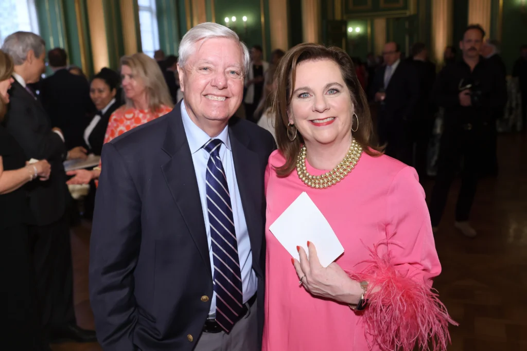 A man in a suit and a woman in a pink dress with feathered sleeves smile at an indoor event, surrounded by other people in formal attire.