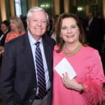 A man in a suit and a woman in a pink dress with feathered sleeves smile at an indoor event, surrounded by other people in formal attire.