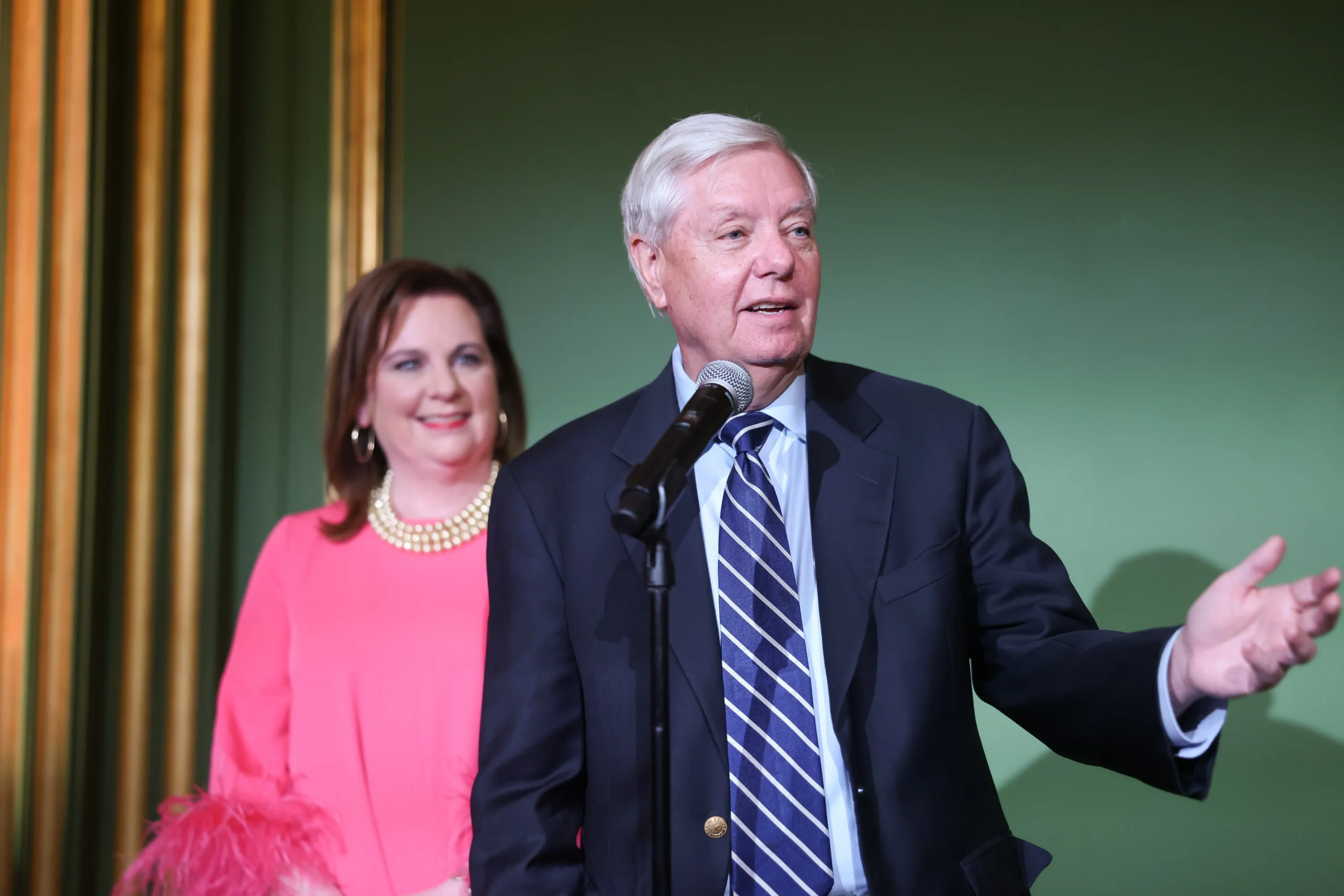 An older man in a suit speaks into a microphone, gesturing with one hand, while a woman in a pink outfit with a pearl necklace stands behind him, smiling. The background is green with a gold frame.