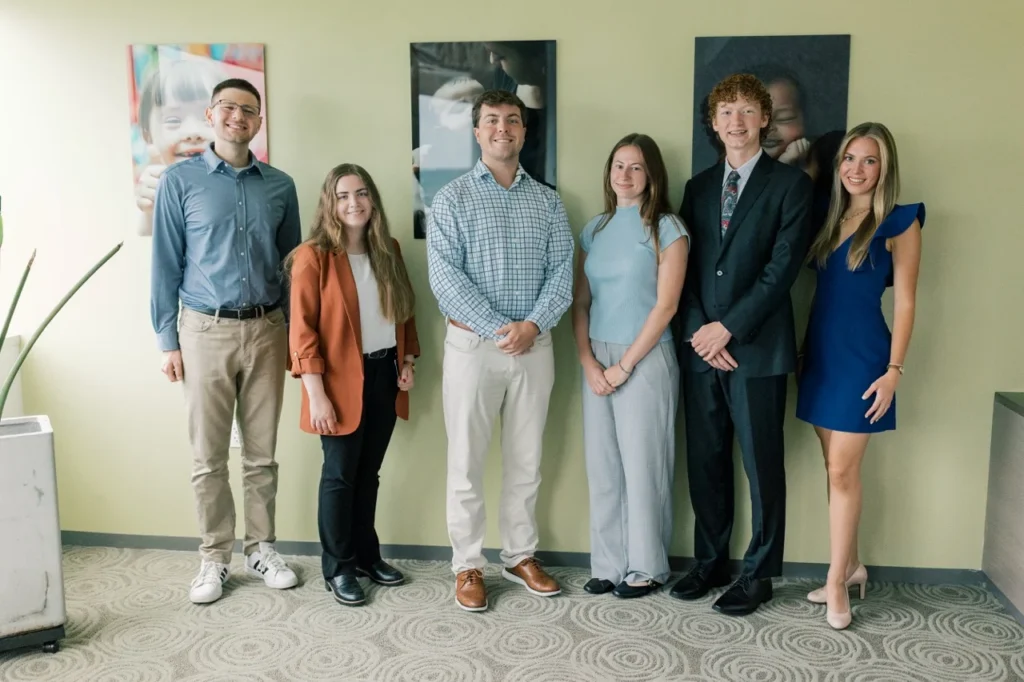 Six people, including SBA Interns, stand side by side in business casual and formal attire, smiling at the camera in an office setting with green walls and framed photos behind them—a snapshot of professional life thriving despite today’s dynamic political climate.