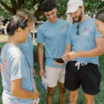 Four people wearing matching blue shirts stand together outdoors, smiling and looking at a phone held by one of them. As part of Parker's Journey as a Student Canvasser, they appear happy and are gathered under a tree on a sunny day.