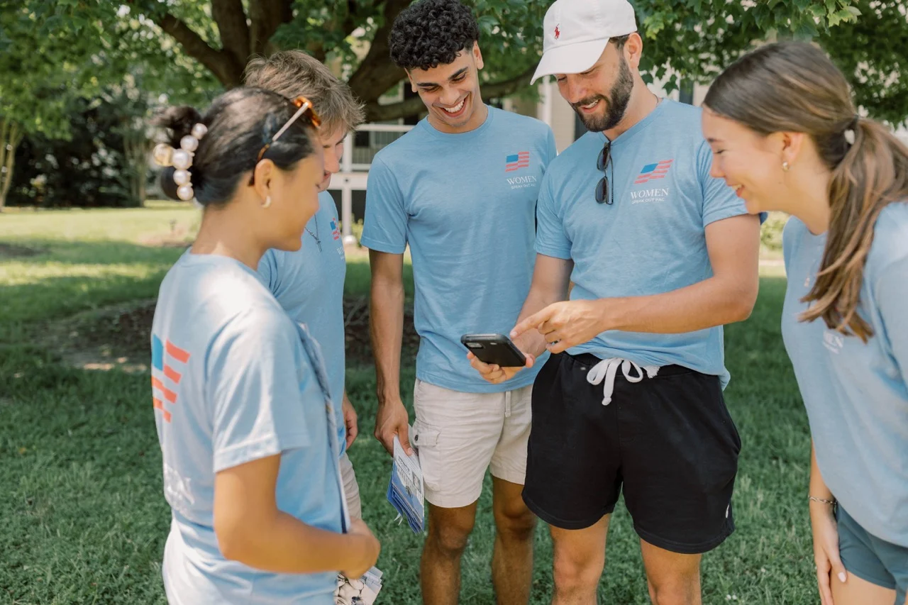 Four people wearing matching blue shirts stand together outdoors, smiling and looking at a phone held by one of them. As part of Parker's Journey as a Student Canvasser, they appear happy and are gathered under a tree on a sunny day.