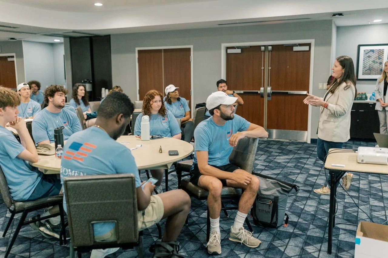 A group of young adults in blue shirts sit around tables in a conference room, listening to a woman—herself a full-time pro-lifer—standing and speaking at the front. Some take notes, while others look attentive, inspired by stories like Parker’s journey as a student canvasser.