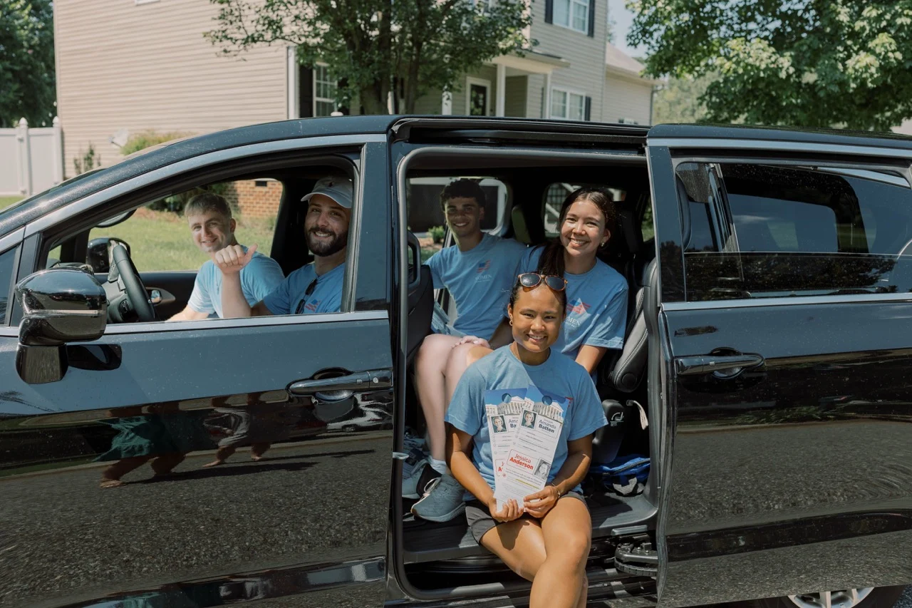 Five smiling people in blue shirts, including Parker—the full-time pro-lifer and enthusiastic student canvasser—sit in and beside a black van with open doors. The person in front holds flyers. Houses and trees are visible in the background on a sunny day.