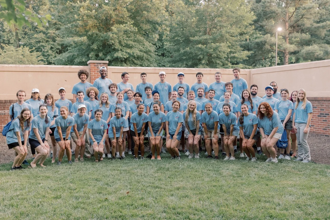 A large group of people in matching blue shirts and shorts, including a dedicated Student Canvasser from Parker’s Journey, pose together outside on grass, with trees and a brick wall in the background.