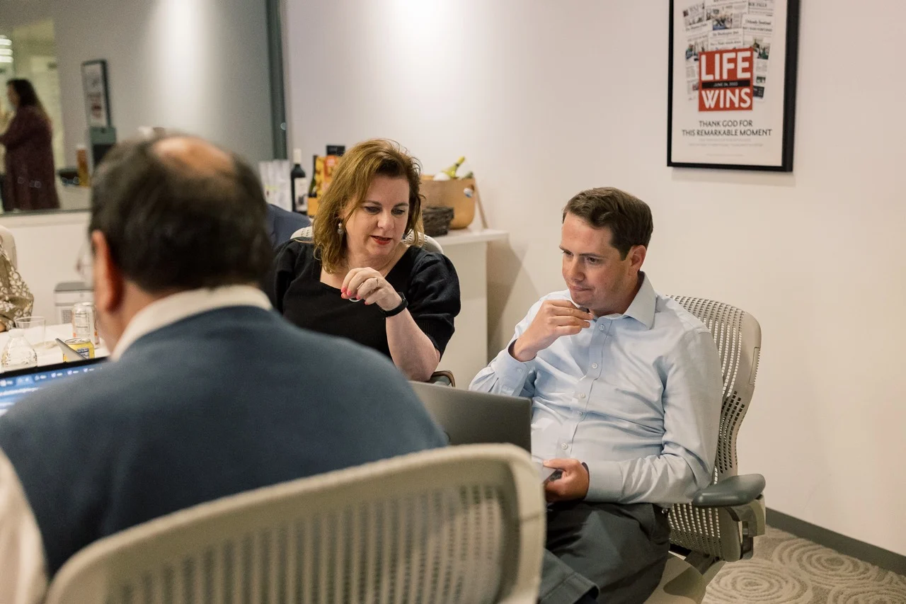 Two people, possibly interns or pro-life leaders, sit in office chairs looking at a laptop while another person sits nearby. A framed magazine cover on the wall reads “LIFE WINS.” The setting appears to be a modern office or conference room, perhaps associated with SBA.