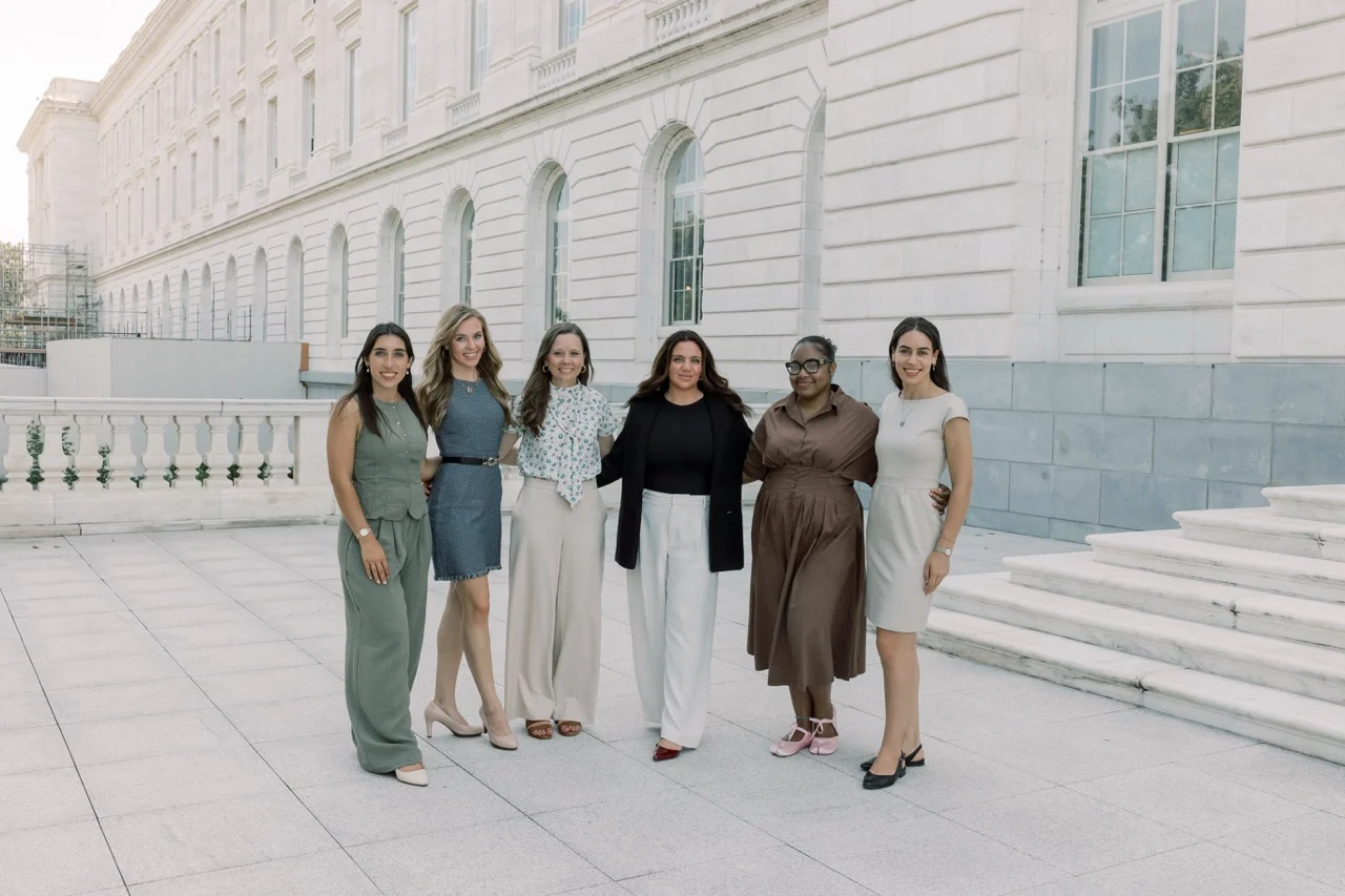 Six women, all SBA Interns, stand together outside a white stone building with large windows, dressed in various business casual outfits and smiling toward the camera on a bright day. Their unity and enthusiasm reflect their commitment to making a difference in today’s political climate.