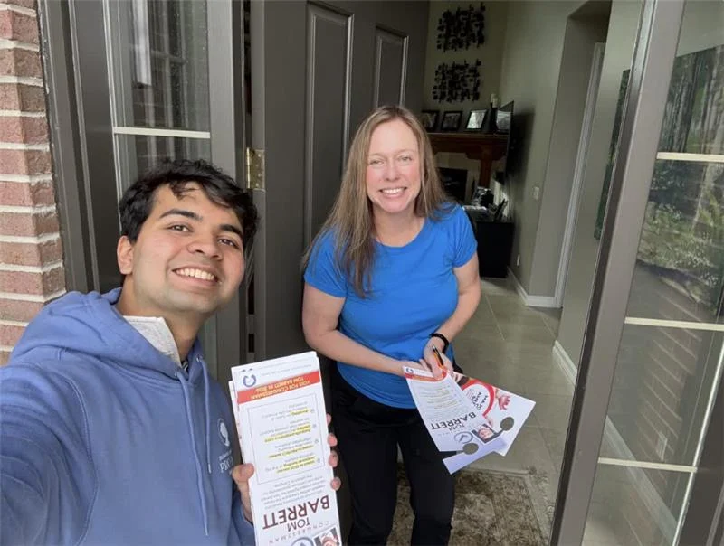 Two people smile at the camera in a doorway. The person on the left, a student canvasser named Parker, holds brochures and wears a blue hoodie; the person on the right, wearing a blue shirt, stands inside the house also holding flyers.