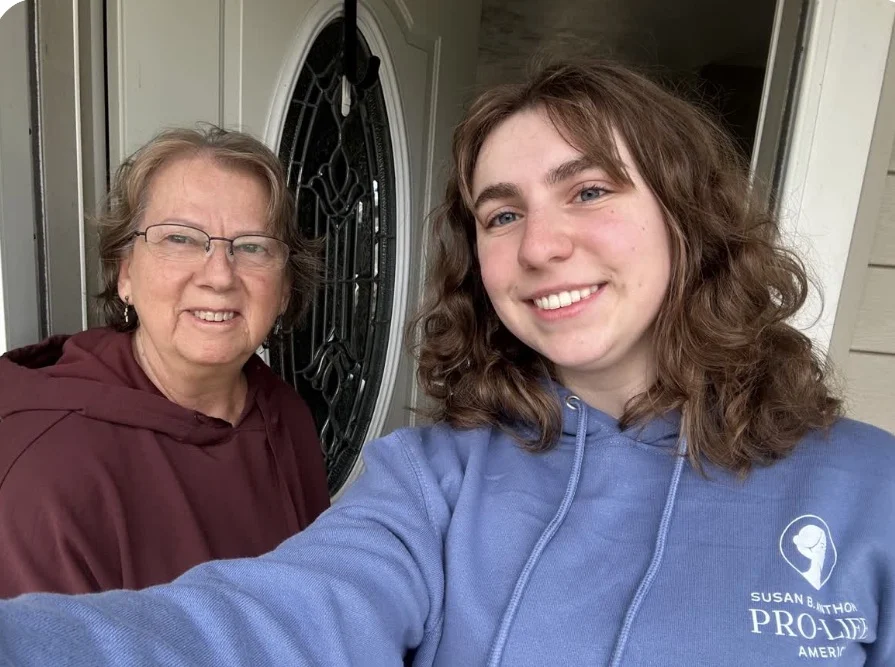 Two people standing in front of a door, smiling for a selfie. One is Parker, wearing glasses and a maroon hoodie, while the other—likely a student canvasser or full-time pro-lifer—has curly hair and sports a blue Susan B. Anthony Pro-Life hoodie.