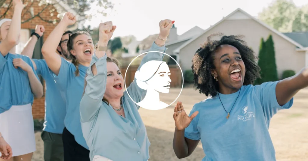 A group of women outdoors cheer and raise their fists, smiling excitedly. Wearing blue shirts emblazoned with a white logo of a woman’s profile centered over the image, these Student Canvassing volunteers radiate energy as they connect with Battleground Voters. Houses are visible in the background, underscoring their commitment to grassroots engagement.