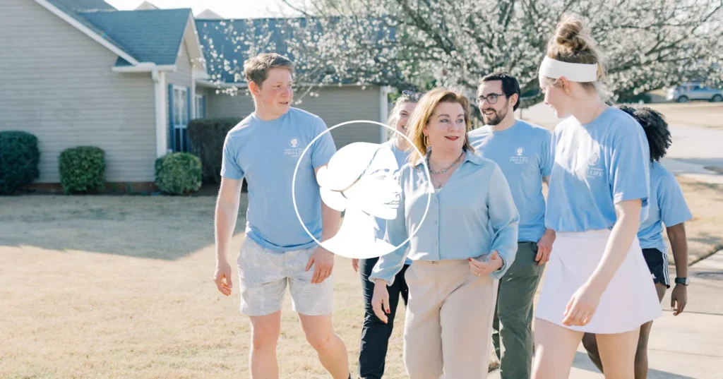 A group of people in casual clothes walk and talk outside in a suburban neighborhood on a sunny day, engaging with battleground voters as part of a Voter Contact Program. A white logo of a woman’s profile is overlaid in the center of the image.