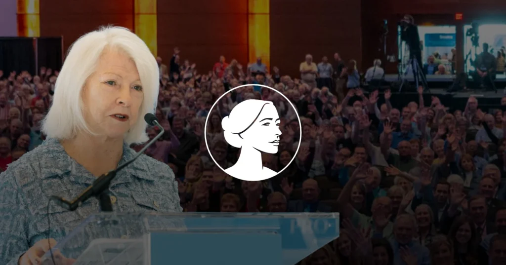 An older woman with white hair, a prominent Pro-Life Leader, speaks at a podium. Behind her, a large audience sits in the hall. There is a white graphic of a woman's profile over the image's center.