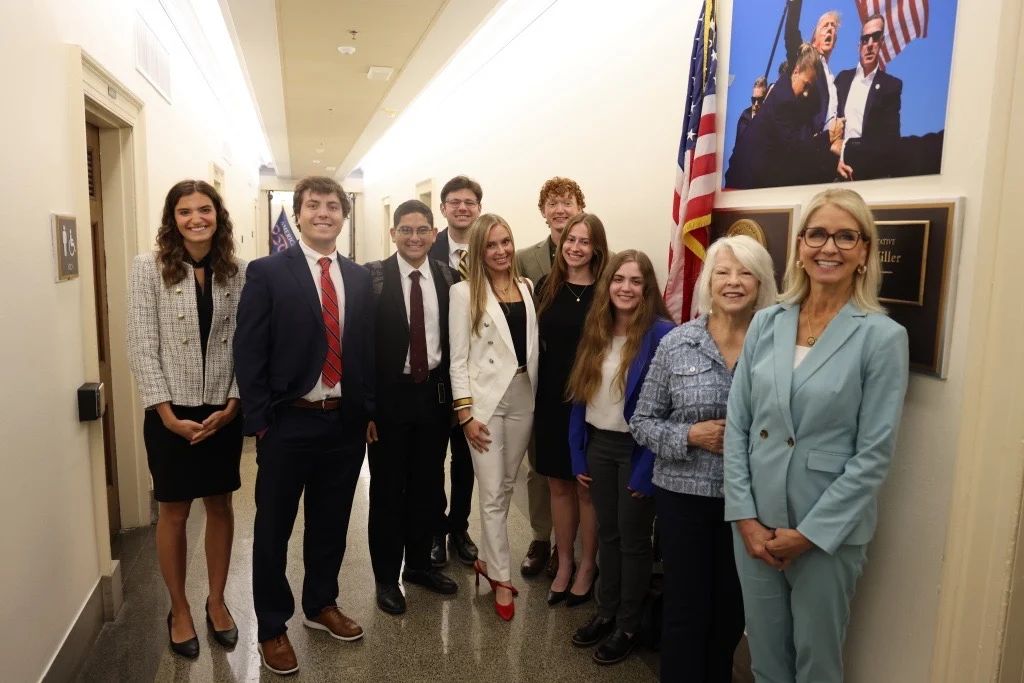 A group of ten people, dressed in business attire, standing and smiling in a hallway near an American flag and a framed photo of leaders—possibly during a Day on the Hill visit with Congresswoman Mary Miller—with office doors visible in the background.
