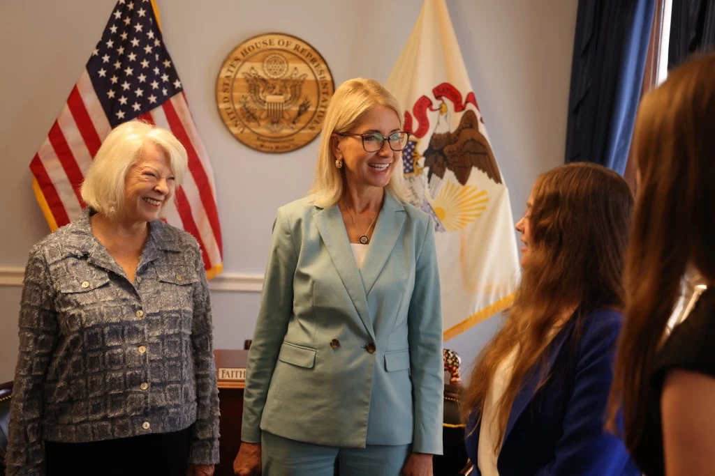 Three women stand and talk in an office with American and Illinois flags, a gold seal on the wall, and a desk in the background. Two women are smiling and wearing business attire, as they participate in Day on the Hill with Congresswoman Mary Miller.