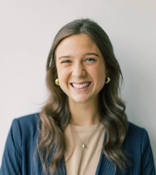 A young woman with long brown hair, wearing a dark blazer and beige top, smiles warmly at the camera against a plain light background—reminiscent of Kathleen Wilber’s poised and approachable presence.