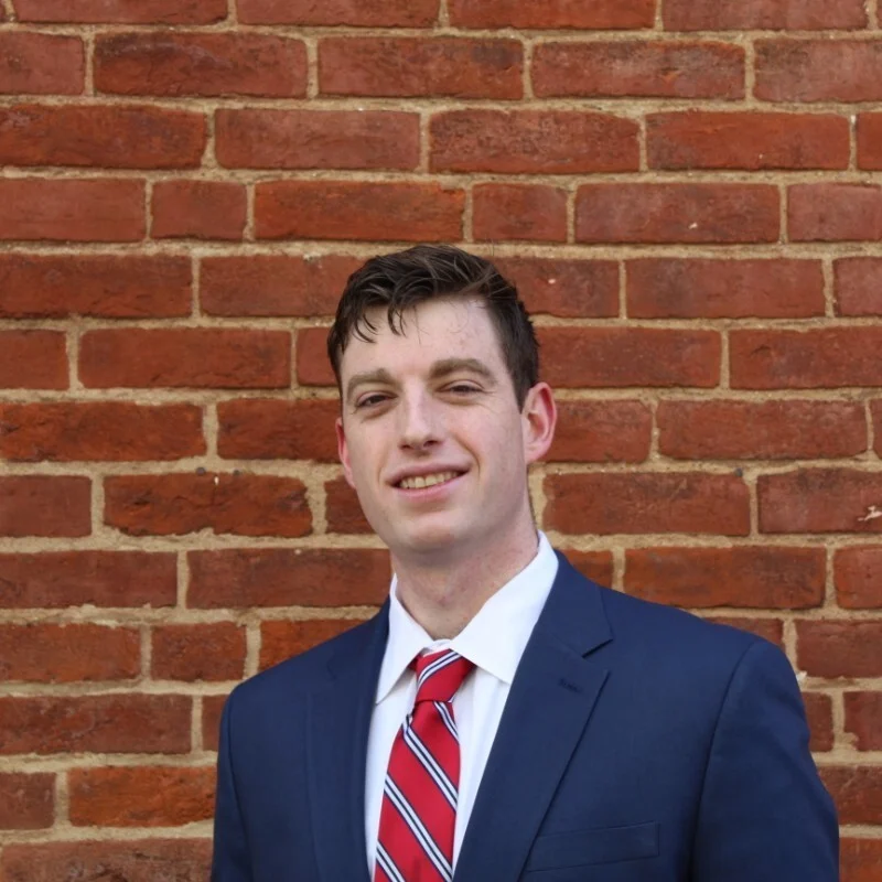 A man in a navy suit, white shirt, and red striped tie stands in front of a red brick wall, smiling slightly at the camera.