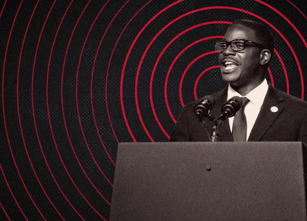 David Crowley in a suit and glasses speaks at a podium against a background of concentric red circles.